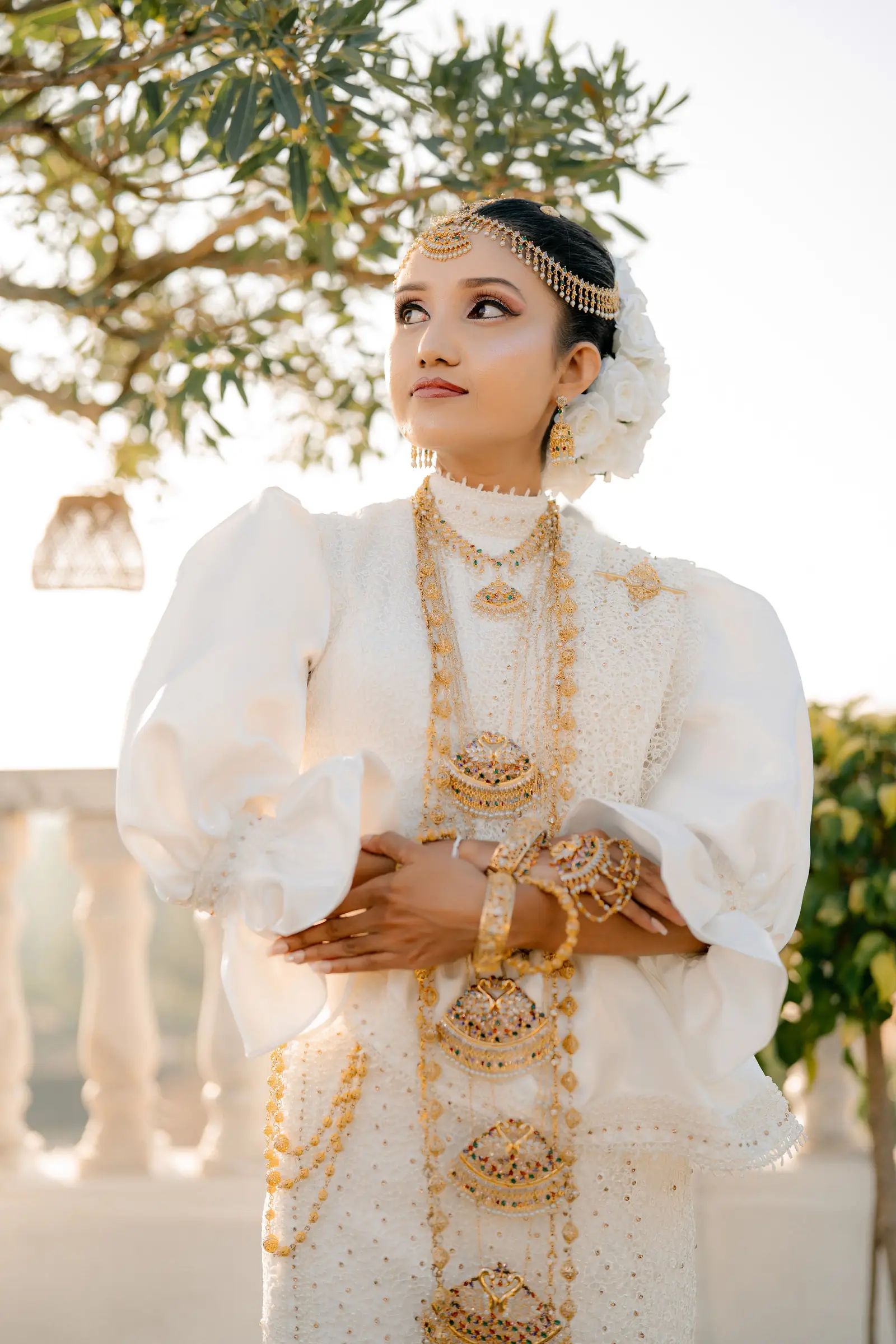 Kandyan bride in white and gold jewellery, hands folded, looking up at golden hour