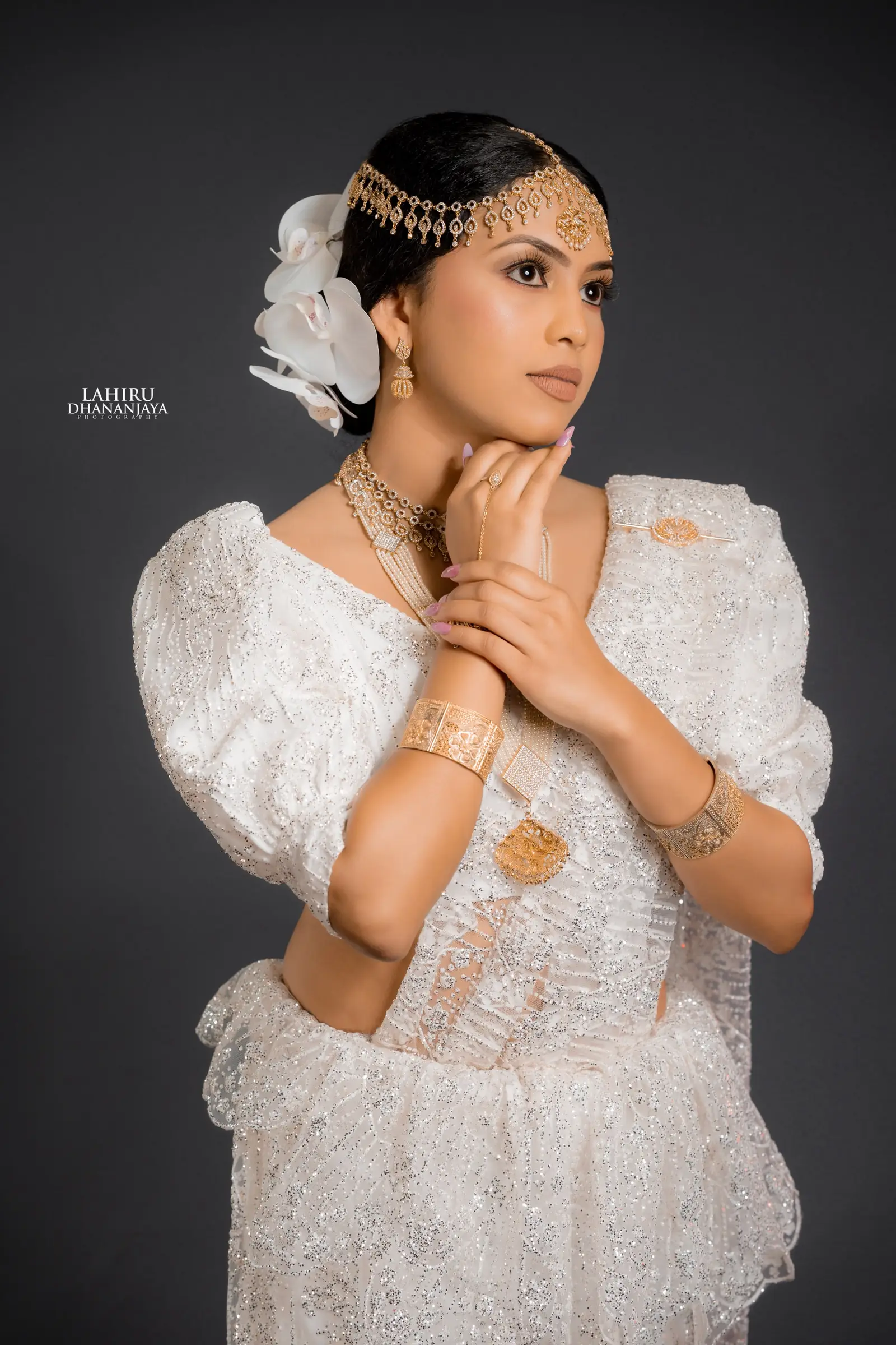 Bride with her hand raised to her chin, wearing gold jewellery on white lace